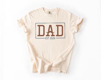 May include: Cream-colored t-shirt with the word "DAD" in brown block letters inside a blue rectangle. A brown mustache graphic is above "EST 2026". The shirt is folded, showing the sleeves and collar.