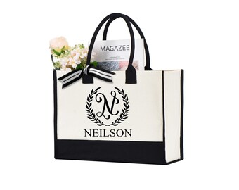 May include: A white canvas tote bag with black trim and a black and white monogram design. The monogram features a stylized letter "N" inside a wreath. The text "NEILSON" is printed below the monogram. The bag is shown with a magazine and flowers inside.
