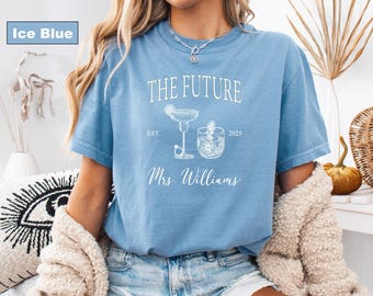 May include: Ice blue t-shirt with the text "THE FUTURE" and a cocktail graphic. Below the graphic is "Mrs. Williams" and the year "2025". The shirt is a light blue color.