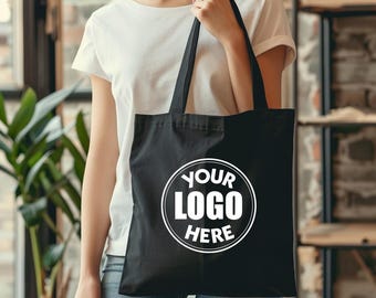 May include: Black tote bag with white circular logo that says "YOUR LOGO HERE". The bag is held by a person wearing a white t-shirt and blue jeans. The background includes a window and a plant.