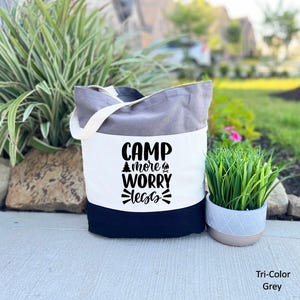 May include: A tri-color grey tote bag with the text "CAMP more WORRY less" in black. The bag has a white body, a grey top, and a navy blue bottom. It has a white handle and is sitting on a concrete surface.