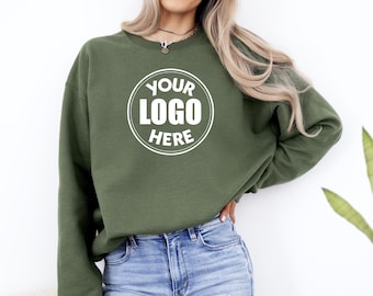 May include: Olive green sweatshirt with the text "YOUR LOGO HERE" in a white circle. The sweatshirt is a crew neck style and is made of a soft material. The model is wearing blue jeans.