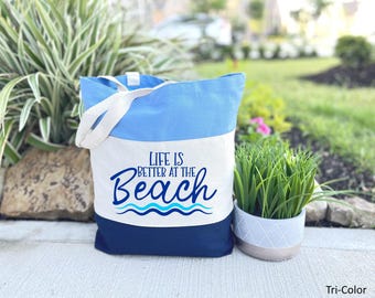 May include: A blue and white canvas tote bag with the text "Life is Better at the Beach" in blue and white lettering. The bag has a blue and white wave design and blue handles.