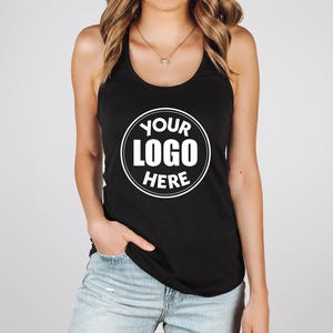 May include: Black racerback tank top with a white circular logo that reads "YOUR LOGO HERE". The tank top is sleeveless and has a scoop neck. The model is wearing blue jeans.