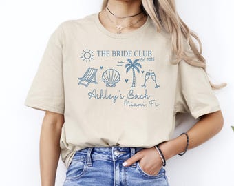 May include: Beige t-shirt with a blue beach-themed graphic. The design includes a sun, beach chair, seashell, palm tree, champagne flutes, and the text "THE BRIDE CLUB Est. 2025" and "Ashley's Bach Miami, FL".