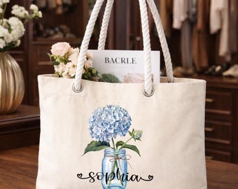 Peut inclure: Un sac fourre-tout en toile couleur crème avec des poignées en corde blanche et des œillets argentés. L'avant présente un motif d'hortensia bleu dans un bocal en verre avec le nom Sophia imprimé en lettres cursives noires sous le bocal.