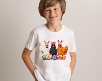 Peut inclure: T-shirt blanc avec trois poulets brodés portant des oreilles de lapin, debout sur des œufs de Pâques décorés. Les poulets sont blancs, noirs et orange. Les œufs sont colorés. Le t-shirt est porté par un enfant.