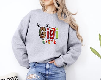 Peut inclure: Un sweat-shirt gris clair avec le mot "Gigi" dans un design de Noël festif. La lettre "G" est décorée d'un motif marron et vert, et les lettres "i" sont rouges et vertes. Une guirlande de bas de Noël est suspendue en dessous.