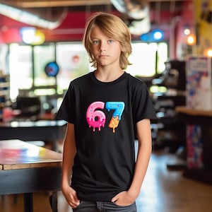 May include: Black t-shirt featuring the number "67" in a colorful, ice cream-themed design. The "6" is pink and donut-shaped, while the "7" is blue with a waffle cone. The shirt is worn by a person.