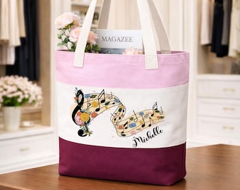 Peut inclure: Un sac fourre-tout en toile à blocs de couleurs avec une section supérieure rose clair, une section centrale blanche ornée d'un motif de notes de musique florales et du nom Michelle, et une base bordeaux foncé avec deux anses en tissu crème.