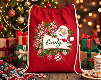 Peut inclure: Un sac à dos à cordon rouge orné d'un motif représentant le Père Noël au-dessus d'un fanion portant le prénom Emily écrit en lettres vertes. Le design comprend un grand nœud à pois rouges et imprimé léopard, des branches de pin et des boules.