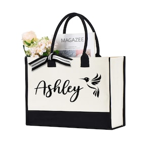 May include: A canvas tote bag with black handles and a black bottom panel. The name "Ashley" is printed in cursive, with a hummingbird graphic. The bag contains flowers and a magazine.