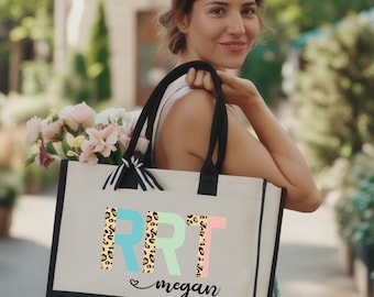 May include: A canvas tote bag with black handles and a black bottom panel. The bag features the letters "RRT" in blue, leopard print, and pink, with the name "megan" below. The bag is filled with pink and white flowers.