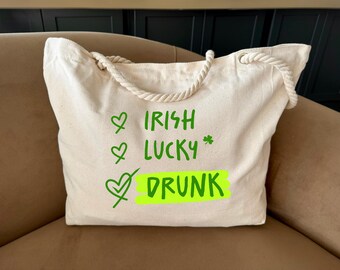 Peut inclure: Sac fourre-tout en toile crème avec des poignées en corde. Le sac présente un texte vert qui dit "Irish, Lucky, Drunk" avec des coches et un trèfle. Le sac est posé sur une surface marron.