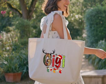 Peut inclure: Un sac fourre-tout crème avec le nom "Gigi" dans un design festif avec des bois de rennes, des bas de Noël et un bonnet de Père Noël. Le sac est porté par une personne dans une robe à fleurs blanche.