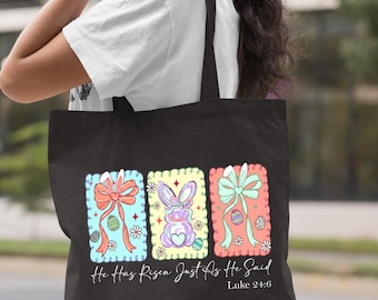 Peut inclure: Sac fourre-tout noir avec trois motifs de Pâques colorés. Chaque motif comprend un nœud, des œufs de Pâques et un lapin. Le texte "He Has Risen Just As He Said Luke 24:6" est imprimé en dessous.