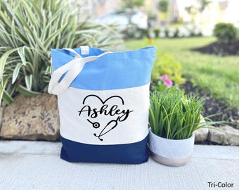 May include: A blue, white, and navy blue tote bag with a heart design and the name "Ashley" in black text. The heart design includes a stethoscope and a medical symbol.