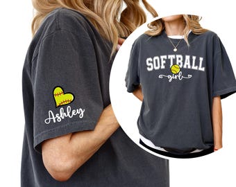May include: Dark gray t-shirt with the text "SOFTBALL girl" and a softball graphic. The left sleeve features a yellow softball heart and the name "Ashley" in script. The shirt is a casual, sporty style.