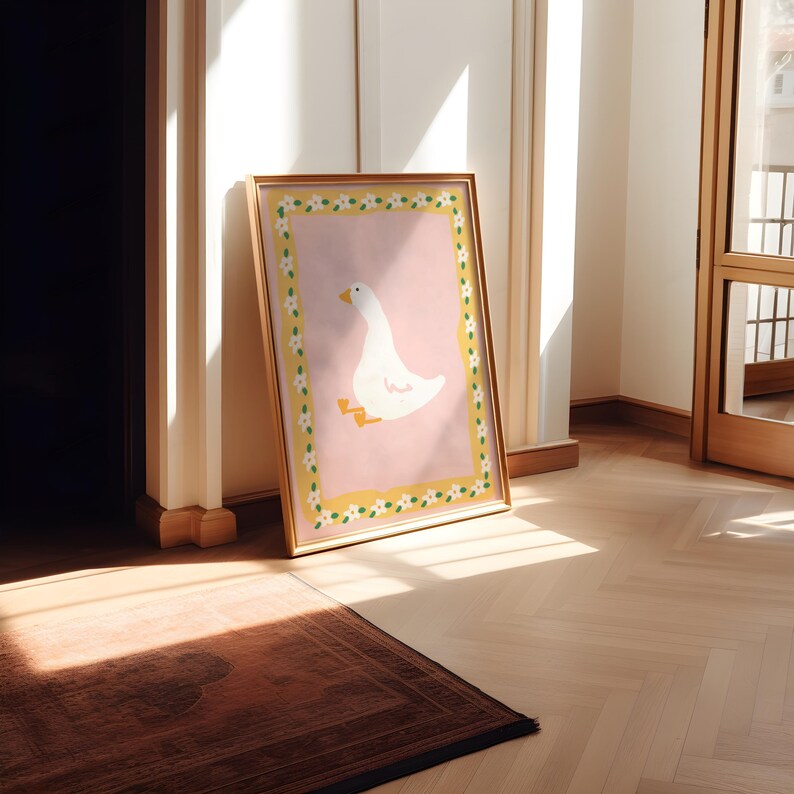 May include: A framed art print featuring a white goose illustration on a pink background. The artwork is bordered by a yellow frame with a floral pattern. The print is leaning against a wall in a room with hardwood floors.
