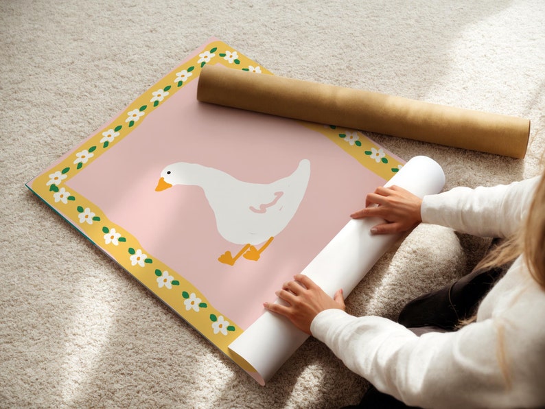 May include: A pink poster with a white goose illustration, yellow border with white flowers, and a brown rolled-up poster. The poster is on a light-colored rug.