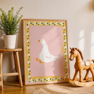 May include: A framed art print featuring a white goose illustration on a pink background, framed with a yellow border adorned with white flowers. A wooden rocking horse and a potted plant are in the foreground, creating a nursery theme.