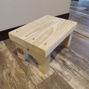 May include: A simple wooden step stool made from unfinished pine wood. The stool has a rectangular top and two legs that are slightly curved.