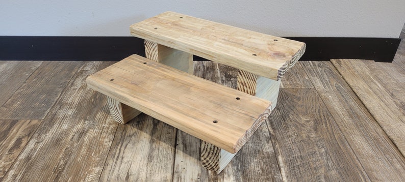 May include: A two-tiered wooden step stool featuring a natural light brown wood grain finish. The structure consists of two rectangular platforms supported by vertical wooden legs, with visible screw holes on the top surfaces of each step.