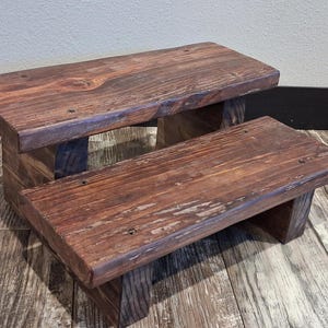 May include: A two-step wooden stool featuring a dark brown stained finish with visible wood grain patterns. The structure consists of two rectangular platforms supported by sturdy vertical wooden legs, creating a tiered design.