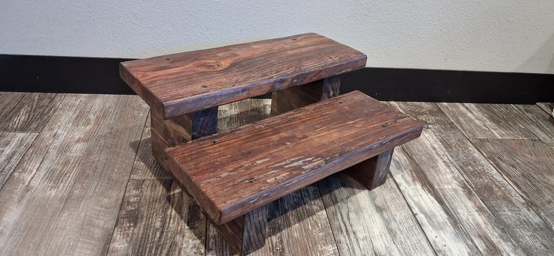 May include: A two-step wooden stool featuring a dark brown stained finish with visible wood grain patterns. The structure consists of two rectangular platforms supported by sturdy vertical wooden legs, creating a tiered design.