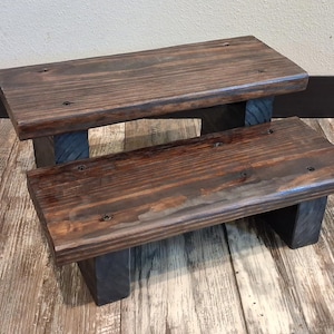 May include: A two-step wooden stool with a dark stain. The steps are rectangular and supported by dark-colored legs. The wood grain is visible, giving it a rustic appearance. This step stool is suitable for various uses.