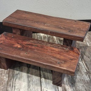 May include: A two-tier wooden step stool featuring a dark brown stained finish with visible wood grain patterns. The structure consists of two rectangular platforms supported by sturdy block legs, with visible screw heads on the top surface.