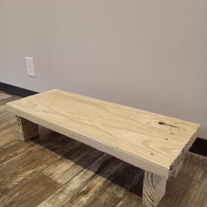 May include: A rectangular wooden step stool with a natural finish. The stool has a flat top and four sturdy legs. The wood grain is visible, and the stool appears to be handmade. The step stool is designed for use in a variety of settings.