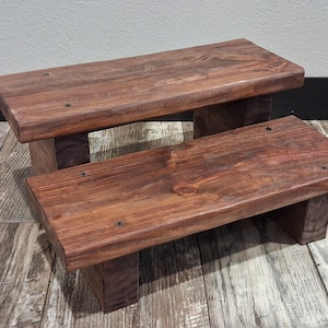 May include: A two-tier wooden step stool featuring a dark brown stained finish with visible wood grain patterns. The structure consists of two rectangular platforms supported by sturdy block legs, showcasing a simple and functional design.