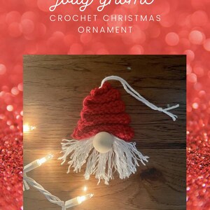 May include: A crocheted gnome ornament with a red hat, white beard, and a wooden nose. The ornament is hanging from a white string and is on a wooden surface with white Christmas lights.