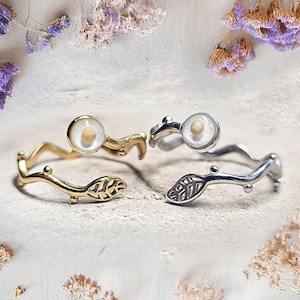 Puede incluir: Cuatro anillos con un diseño inspirado en la naturaleza. Dos son dorados y dos plateados. Cada anillo presenta un engaste claro y circular con un pequeño objeto de color claro en su interior. Las bandas tienen un diseño ondulado en forma de rama con detalles de hojas.