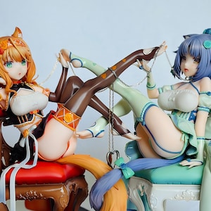 May include: Two anime figurines of female characters, one with orange hair and a fox tail, the other with blue hair and a cat tail, both wearing elaborate outfits and sitting on chairs connected by a chain.