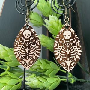 May include: A pair of wooden earrings with a skeleton and floral design. The earrings are brown with white accents and have a teardrop shape.