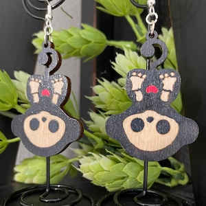 May include: A pair of wooden earrings featuring a black and brown ghost design with red feet. The earrings are hanging from silver hooks and are displayed on black wire stands.