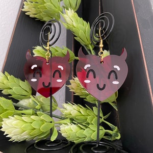 May include: A pair of red heart-shaped earrings with devil horns and a smiling face. The earrings are on a black stand with a gold hook.