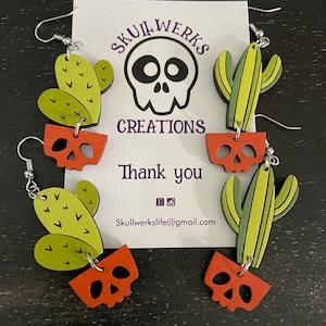 May include: A pair of dangle earrings featuring a green cactus with a red skull pot. The earrings are handmade and have a whimsical, spooky style. The card in the background says "Skullwerks Creations" and "Thank you".
