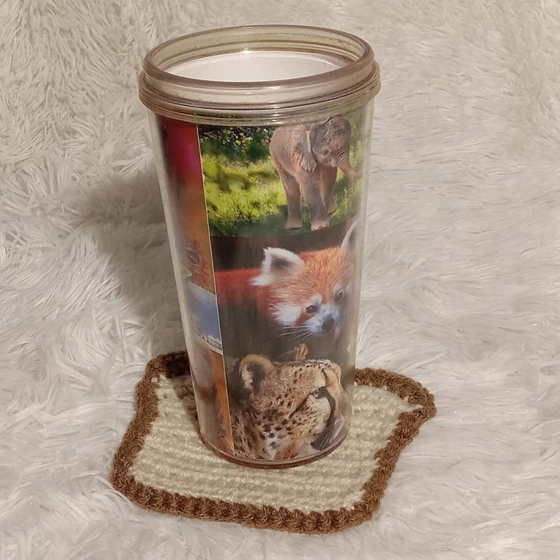 K&ouml;nnte beinhalten: Ein durchsichtiger Plastikbecher mit einem braunen, wei&szlig;en und roten Tierdruckdesign. Der Becher zeigt einen Roten Panda, einen Geparden und einen Elefanten. Der Becher steht auf einem geh&auml;kelten Untersetzer in Braun und Beige.