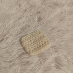 K&ouml;nnte beinhalten: Ein kleiner, quadratischer, geh&auml;kelter Untersetzer in Cremefarbe. Der Untersetzer ist mit einem einfachen Stichmuster gearbeitet und hat eine weiche, strukturierte Oberfl&auml;che.