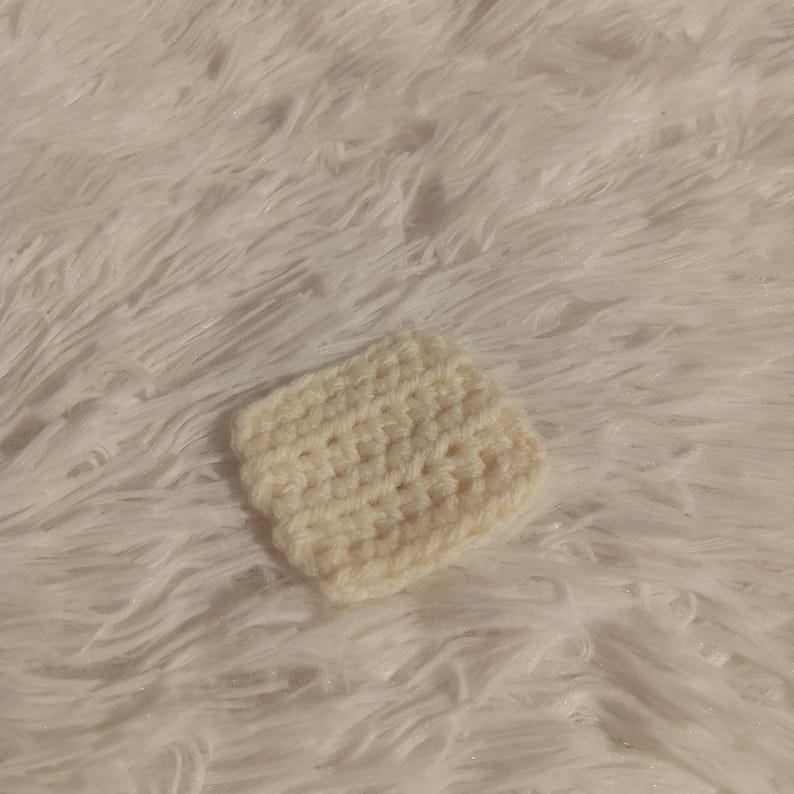 K&ouml;nnte beinhalten: Ein kleiner, quadratischer, geh&auml;kelter Untersetzer in Cremefarbe. Der Untersetzer ist mit einem einfachen Stichmuster gearbeitet und hat eine weiche, strukturierte Oberfl&auml;che.