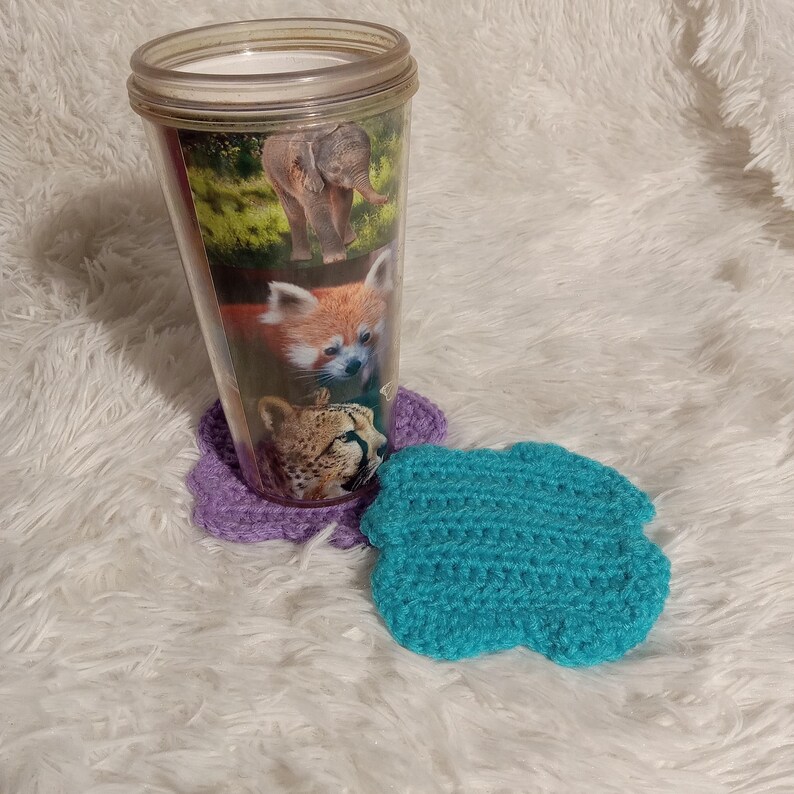 K&ouml;nnte beinhalten: Ein durchsichtiger Plastikbecher mit einem bunten Tierdruckdesign. Der Becher steht auf einem lilafarbenen geh&auml;kelten Untersetzer. Ein blauer geh&auml;kelter Untersetzer befindet sich im Vordergrund.