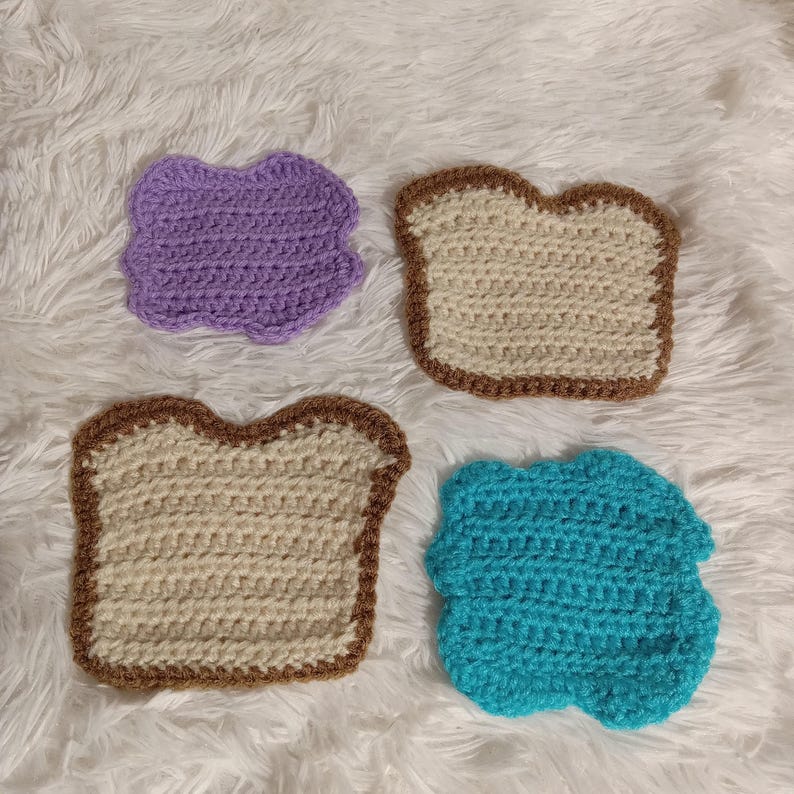 K&ouml;nnte beinhalten: Vier geh&auml;kelte Stoffst&uuml;cke in Form von Brotscheiben. Eine ist lila, eine ist blau und zwei sind beige mit braunen R&auml;ndern.