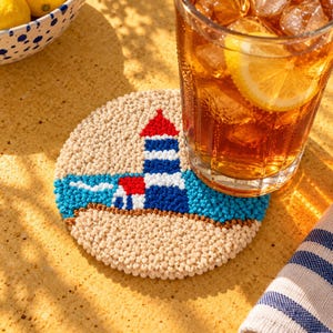 Può includere: Un sottobicchiere rotondo e testurizzato realizzato in filato beige, blu e rosso. Il design presenta un faro con una cima rossa, strisce orizzontali blu e bianche e un mare blu su uno sfondo di sabbia beige, con un bicchiere di tè freddo.