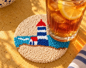 Può includere: Un sottobicchiere rotondo e testurizzato realizzato in filato beige, blu e rosso. Il design presenta un faro con una cima rossa, strisce orizzontali blu e bianche e un mare blu su uno sfondo di sabbia beige, con un bicchiere di tè freddo.