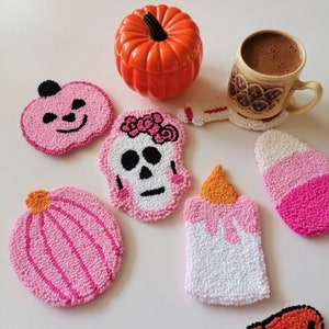 May include: Set of six handmade Halloween-themed coasters featuring a pumpkin, skull, candle, and candy corn designs. The coasters are made with pink, white, and orange yarn and are perfect for adding a festive touch to your home decor.