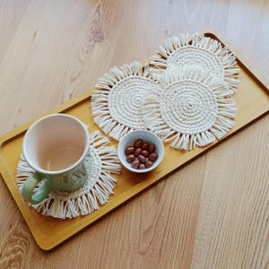 May include: A wooden tray displays a light green mug, a small white bowl of chocolate sweets, and four white macrame coasters. The coasters have a circular design with fringing. The tray rests on a wooden surface.