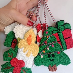 May include: Five handmade Christmas ornaments, each with a different design. The ornaments are made of yarn and feature a Christmas tree, a candle, a present, and two other designs. The ornaments are attached to red and white strings.